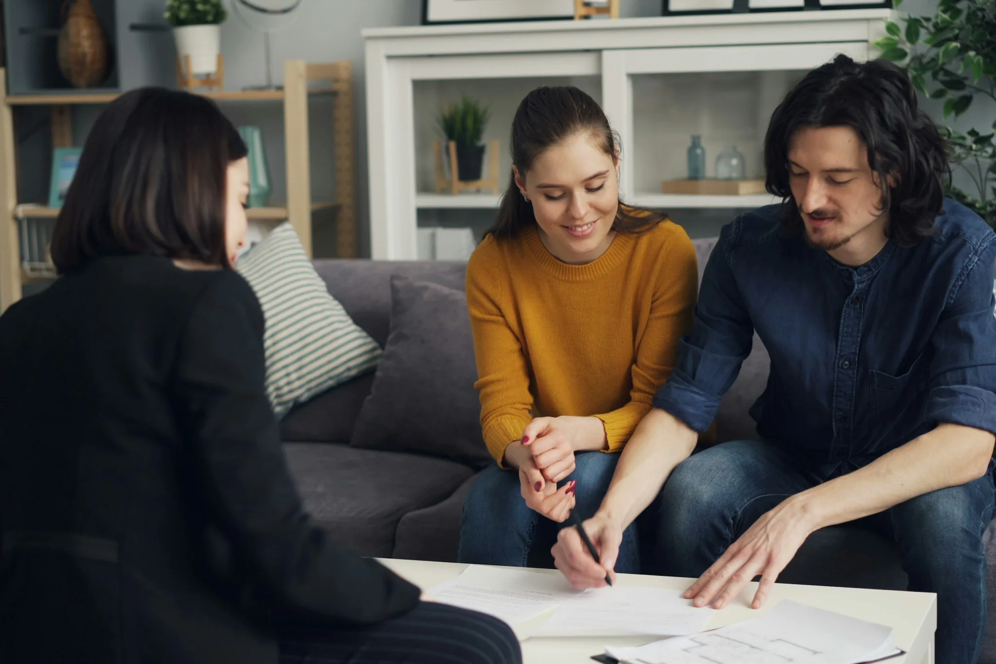 Pareja sentada en un sofá firma los documentos de compra de una vivienda frente a una asesora